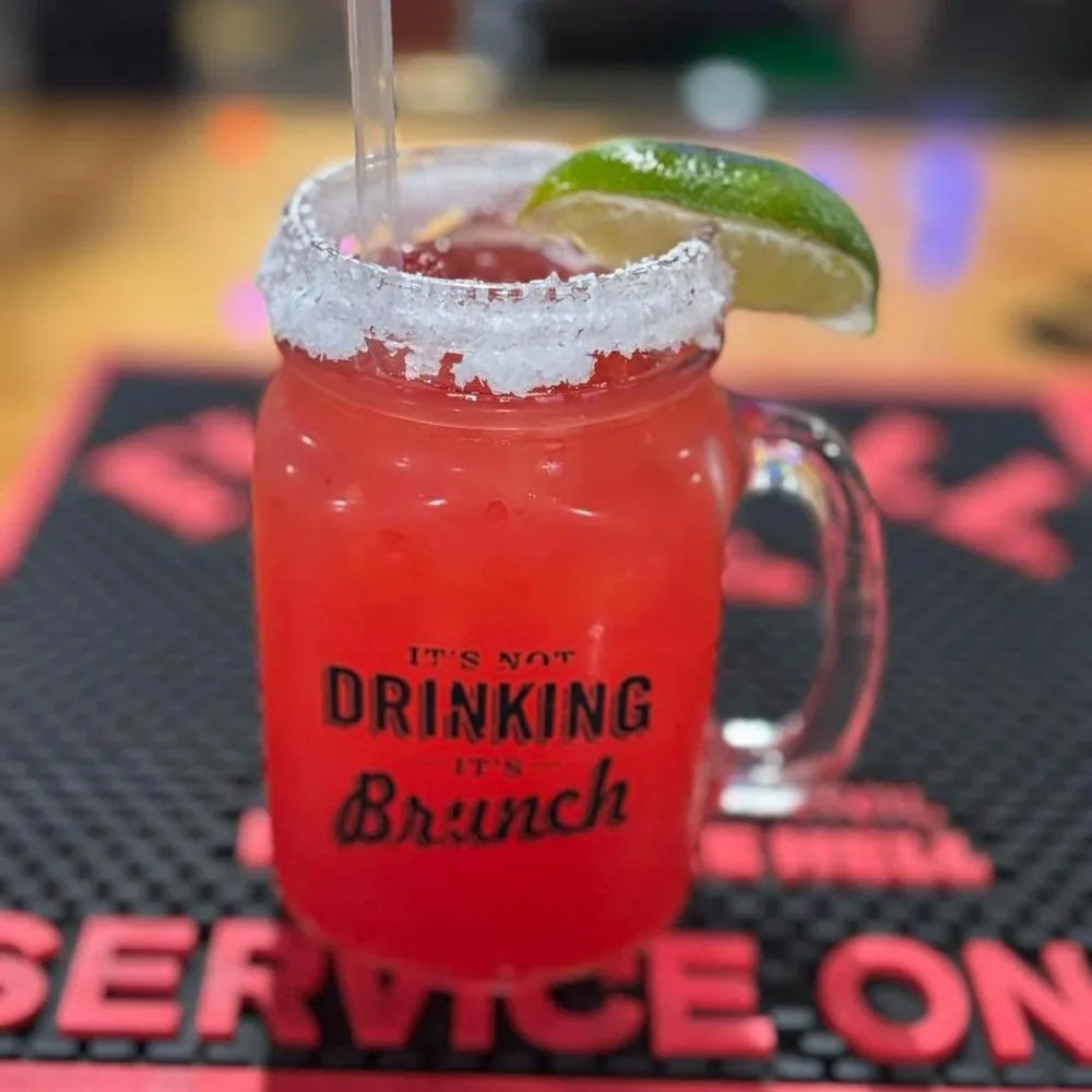A red margarita with a salted rim and a lime wedge in a mason jar mug that reads “It’s not drinking, it’s Brunch”