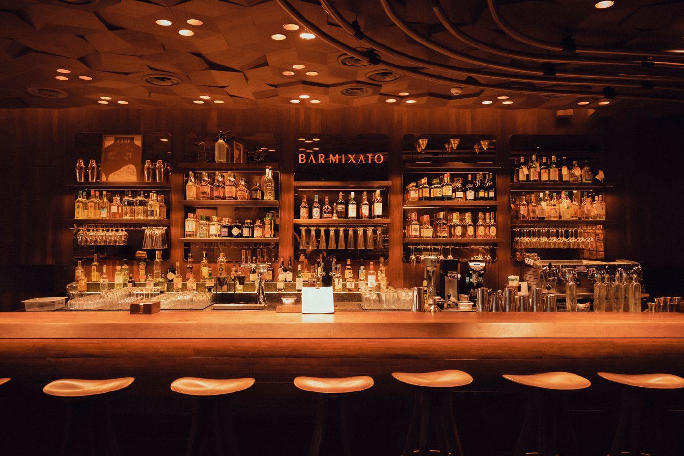 Warm, wood-toned bar interior with shelves of bottles and stools